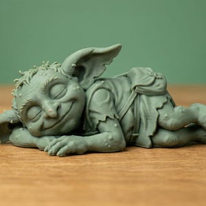 May include: A whimsical, sleeping goblin figurine in a muted green hue. The detailed sculpture features closed eyes, pointed ears, and a gentle smile. The goblin is lying down, with its arms and legs crossed, creating a relaxed pose.