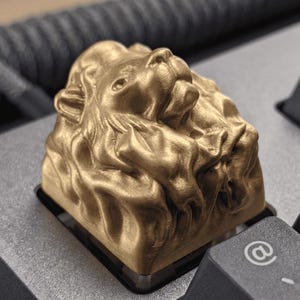 May include: A gold-colored artisan keycap featuring a sculpted lion head with a detailed mane and facial features. The keycap is mounted on a mechanical keyboard next to a black keycap with a white at symbol printed on the surface.