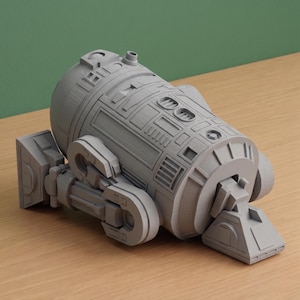 May include: A matte gray 3D printed model of a cylindrical robot featuring intricate mechanical paneling, recessed rectangular vents, circular ports, and two side-mounted leg assemblies with a triangular foot base extending from the front.