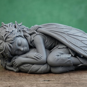Può includere: Statuina di fata grigia in posizione dormiente, con occhi chiusi e braccia incrociate. La fata ha grandi ali dettagliate e riposa su un letto di foglie. La statuina è realizzata in un materiale solido. Sfondo verde.