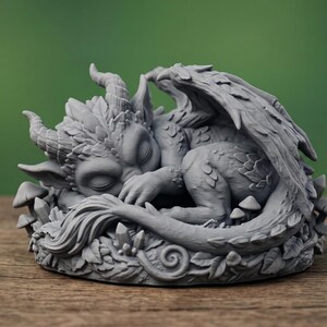 May include: A gray, sleeping dragon figurine with detailed wings, horns, and scales. The dragon is nestled among leaves, mushrooms, and crystals. The sculpture is approximately 4 inches in diameter.