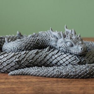 Könnte beinhalten: Graue Drachenfigur, ringförmig auf einer Holzoberfläche liegend. Der Drache hat detaillierte Schuppen, Hörner und einen ruhigen Ausdruck. Der Hintergrund ist einfarbig grün.