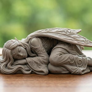 Peut inclure: Une figurine de fée grise en position de sommeil, avec des ailes détaillées et des cheveux fluides. La sculpture repose sur une surface en bois, avec un arrière-plan vert flou. Un carnet, un encrier et une plume sont également visibles.