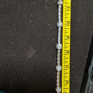 May include: A silver-toned bracelet with five square, crystal-studded beads. The bracelet is measured at approximately 12 inches long with a yellow measuring tape. The background is a dark, textured surface.