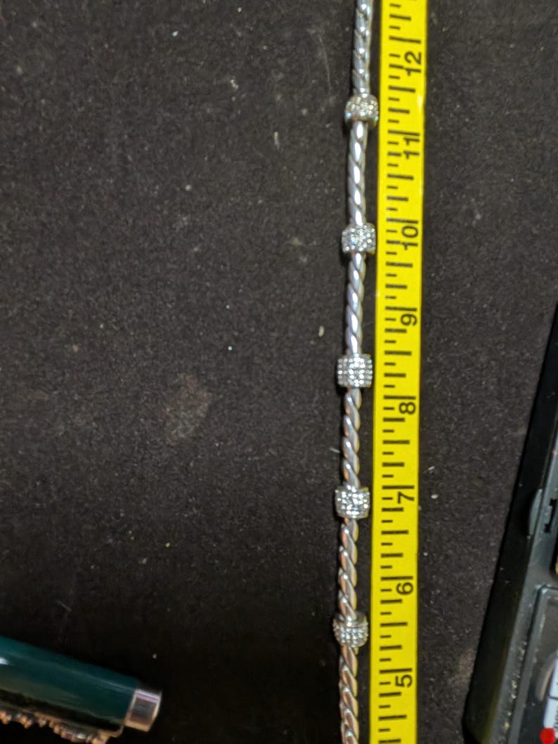 May include: A silver-toned bracelet with five square, crystal-studded beads. The bracelet is measured at approximately 12 inches long with a yellow measuring tape. The background is a dark, textured surface.