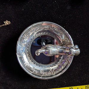May include: A circular silver-toned metal brooch featuring a sculpted cat figure across the centre. The outer rim is decorated with engraved swirling vine patterns and small paw prints. A yellow measuring tape shows the item is 5 cm wide.
