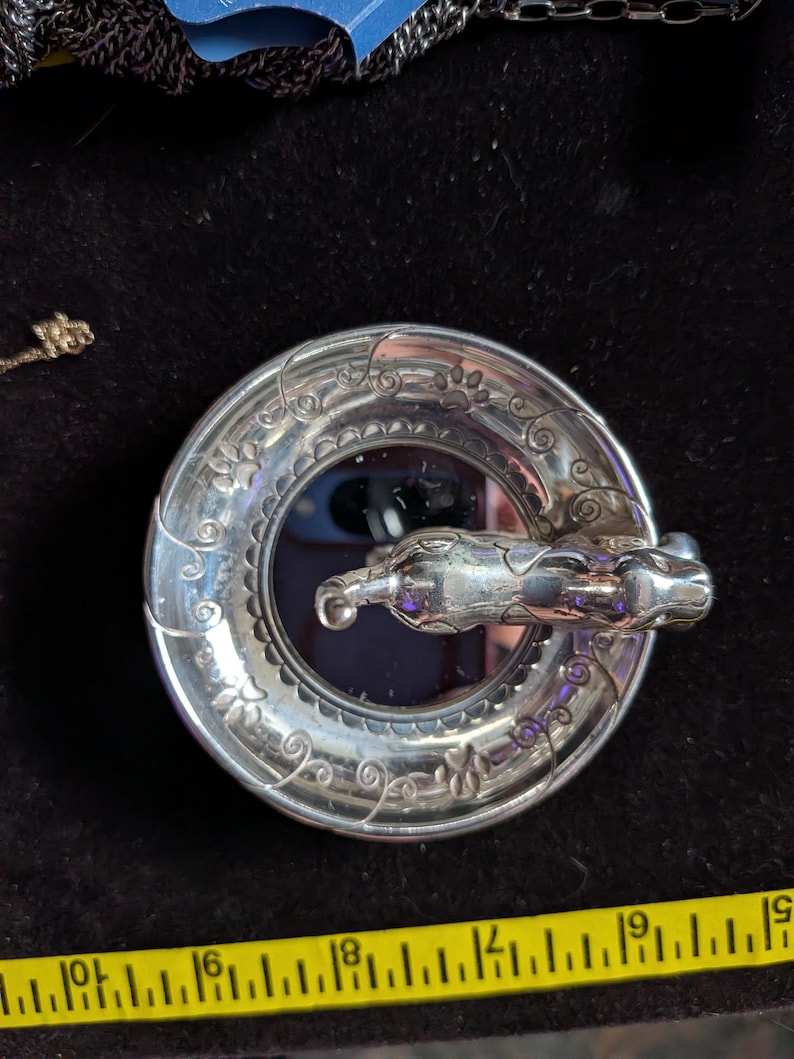 May include: A circular silver-toned metal brooch featuring a sculpted cat figure across the centre. The outer rim is decorated with engraved swirling vine patterns and small paw prints. A yellow measuring tape shows the item is 5 cm wide.
