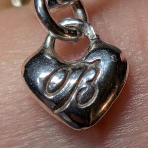 May include: Close-up of a silver heart-shaped charm with a stylized "B" design. The charm is attached to a silver chain link. The charm has a polished, reflective surface.