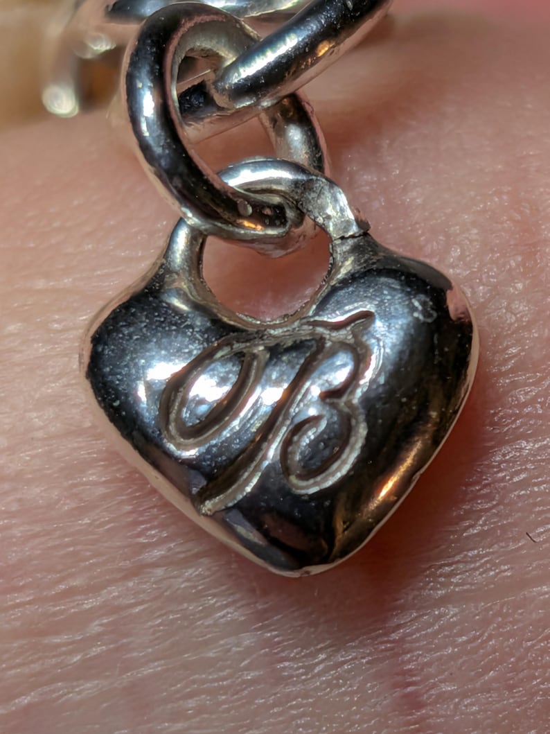May include: Close-up of a silver heart-shaped charm with a stylized "B" design. The charm is attached to a silver chain link. The charm has a polished, reflective surface.