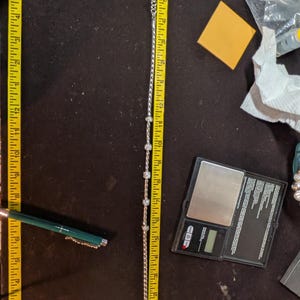 May include: A silver-toned bracelet with clear, round embellishments, measuring approximately 10 inches in length. The bracelet is laid out on a dark surface next to a measuring tape. A pen and a small scale are also visible.