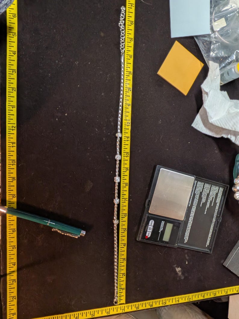 May include: A silver-toned bracelet with clear, round embellishments, measuring approximately 10 inches in length. The bracelet is laid out on a dark surface next to a measuring tape. A pen and a small scale are also visible.