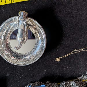 May include: A silver-toned circular trinket dish featuring a sculpted cat figure in the centre and an embossed floral vine pattern on the rim. A yellow measuring tape is visible at the top, showing the dish is approximately 9 cm in diameter.