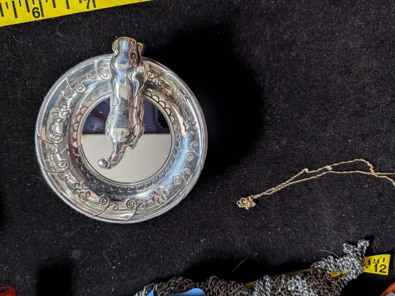 May include: A silver-toned circular trinket dish featuring a sculpted cat figure in the centre and an embossed floral vine pattern on the rim. A yellow measuring tape is visible at the top, showing the dish is approximately 9 cm in diameter.