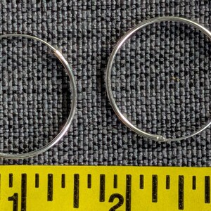 May include: Two thin, circular silver hoop earrings are positioned above a yellow measuring tape. The tape shows the earrings have a diameter of approximately 1 inch each. The hoops are made of a smooth, polished silver metal with a simple wire closure.