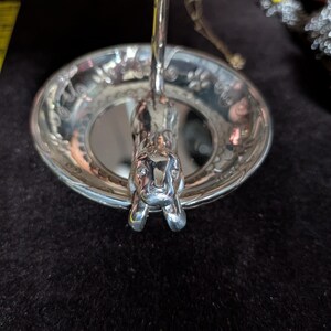 May include: A silver-toned metal trinket dish featuring a sculpted cat figure extending from the centre. The circular bowl has a decorative rim with embossed paw prints. A yellow measuring tape is visible to the left, showing a length of 20 cm.