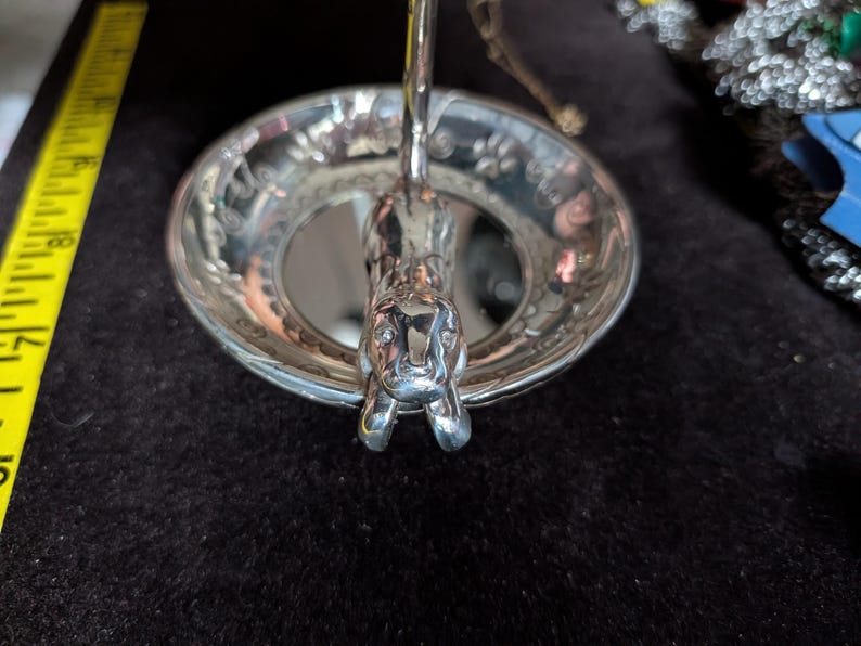 May include: A silver-toned metal trinket dish featuring a sculpted cat figure extending from the centre. The circular bowl has a decorative rim with embossed paw prints. A yellow measuring tape is visible to the left, showing a length of 20 cm.