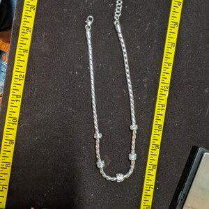 May include: A silver-toned necklace with a twisted rope design and sparkling accents. The necklace is displayed on a dark surface next to a yellow measuring tape. The necklace appears to be approximately 10 inches long.