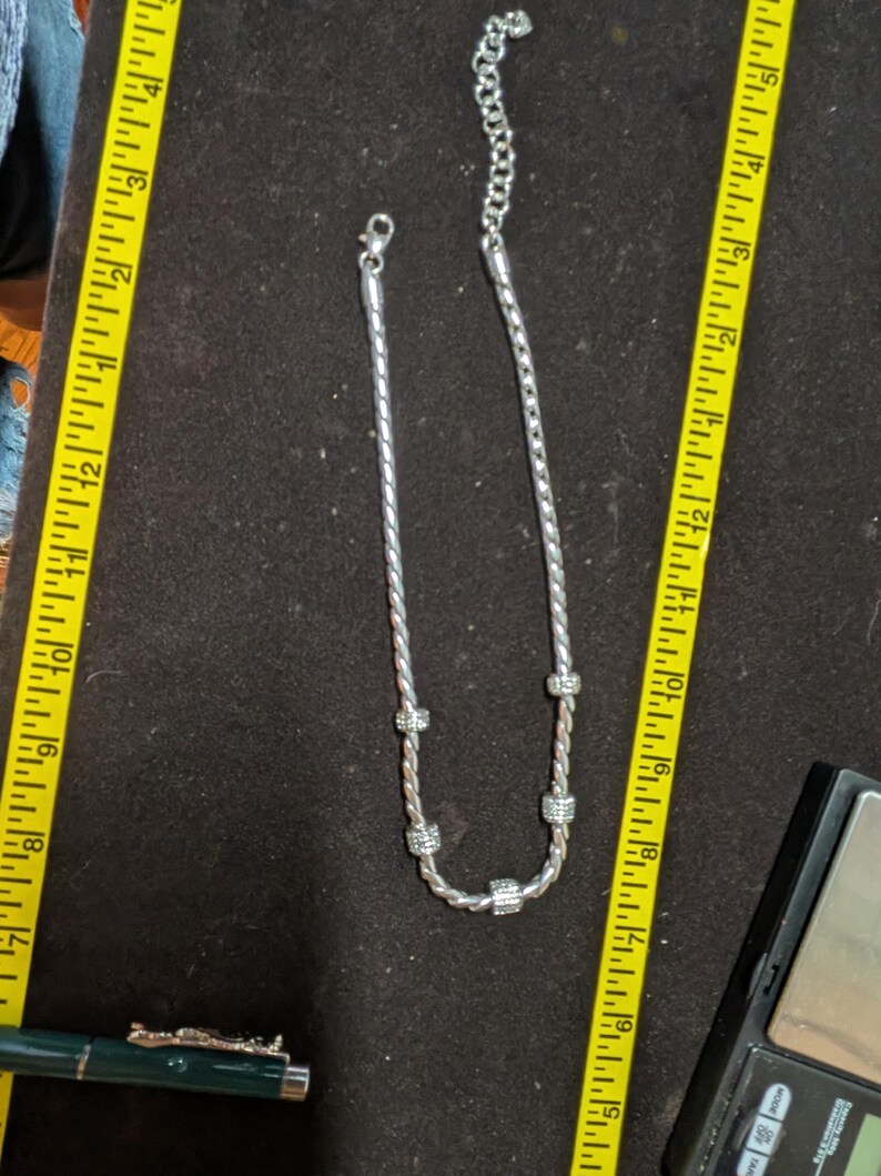 May include: A silver-toned necklace with a twisted rope design and sparkling accents. The necklace is displayed on a dark surface next to a yellow measuring tape. The necklace appears to be approximately 10 inches long.