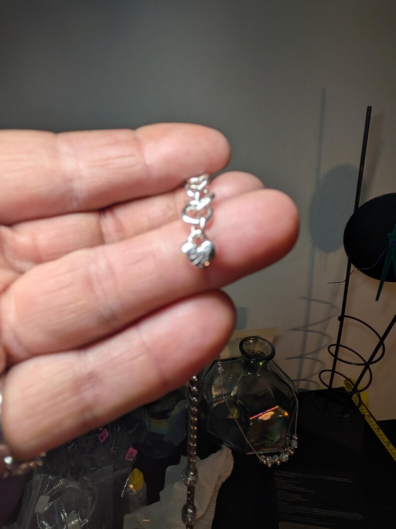May include: A silver-colored chain bracelet with a heart-shaped charm. The charm is suspended from a series of linked heart-shaped links. The bracelet is held in a person's hand.