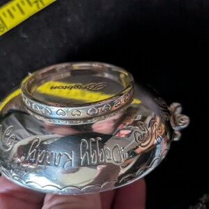 May include: A silver-toned metal bowl with a reflective surface, featuring the engraved text "Doggy Randy" and a decorative heart-patterned rim. A yellow measuring tape is visible, showing a width of approximately 6.35 cm across the top.