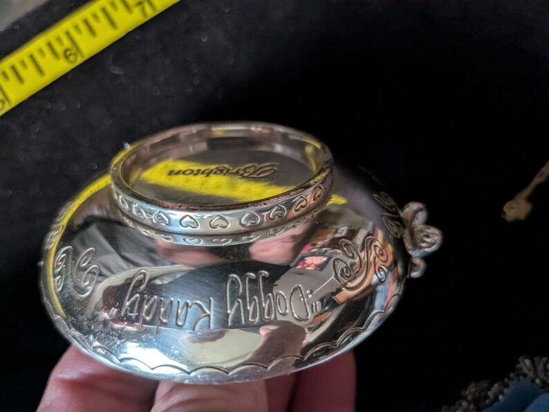 May include: A silver-toned metal bowl with a reflective surface, featuring the engraved text "Doggy Randy" and a decorative heart-patterned rim. A yellow measuring tape is visible, showing a width of approximately 6.35 cm across the top.