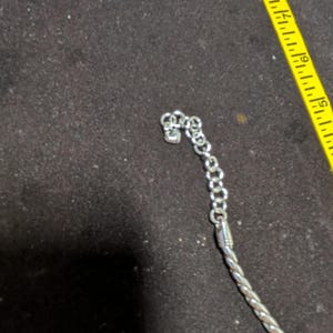 May include: Silver-toned necklace with a braided rope design and a chain clasp. The necklace is partially visible against a dark background, with a yellow measuring tape in the frame.