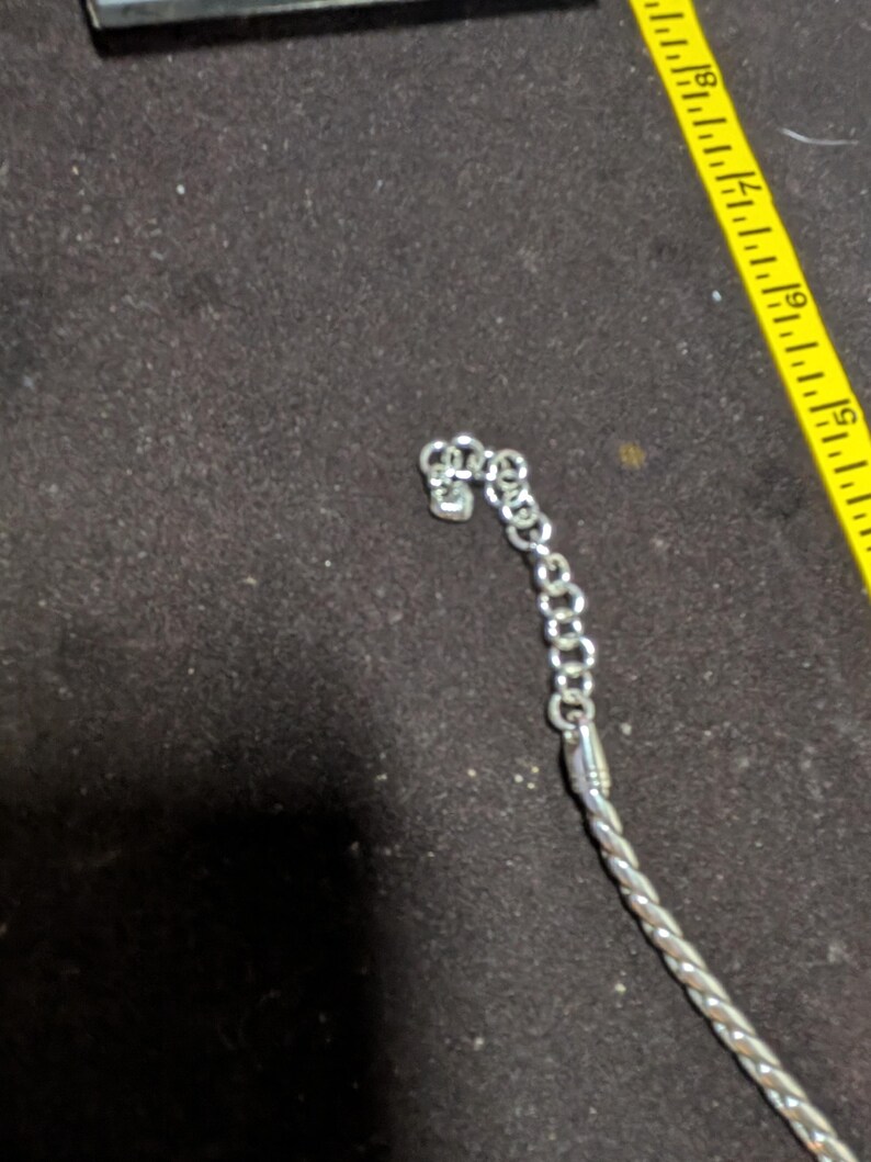 May include: Silver-toned necklace with a braided rope design and a chain clasp. The necklace is partially visible against a dark background, with a yellow measuring tape in the frame.