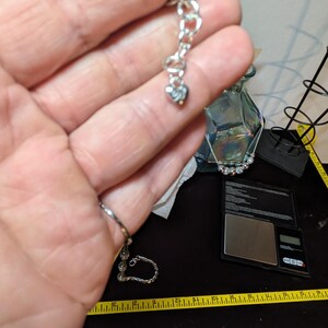 May include: A silver-toned bracelet with a heart-shaped charm. The bracelet is held up against a background that includes a scale, a measuring tape, and a glass vase.