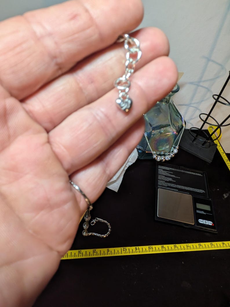 May include: A silver-toned bracelet with a heart-shaped charm. The bracelet is held up against a background that includes a scale, a measuring tape, and a glass vase.