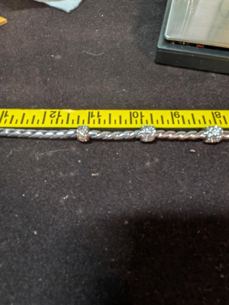 May include: A silver-colored bracelet with three clear, round, decorative beads. The bracelet is measured with a yellow ruler. The beads are approximately 0.25 inches in diameter.