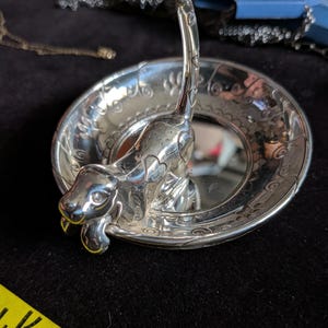 May include: A silver-toned metal ring dish featuring a sculpted dog figure in the centre with an upright tail. The circular dish has an embossed decorative border pattern. A yellow measuring tape is visible, showing a width of approximately 10 cm.