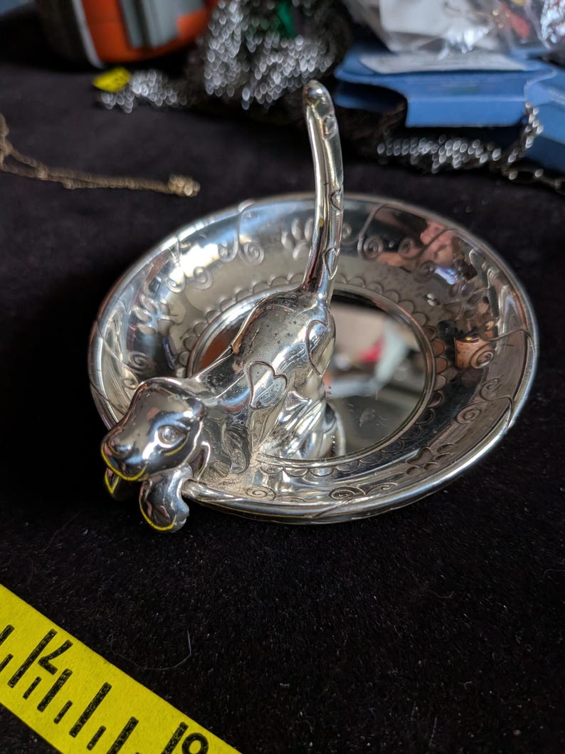 May include: A silver-toned metal ring dish featuring a sculpted dog figure in the centre with an upright tail. The circular dish has an embossed decorative border pattern. A yellow measuring tape is visible, showing a width of approximately 10 cm.