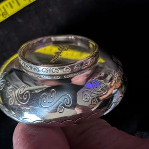 May include: A silver-toned metal bowl with a polished finish and engraved swirl patterns along the sides. A matching silver-toned ring with a heart-patterned band and the word Brighton engraved on the inner rim rests on top of the bowl.
