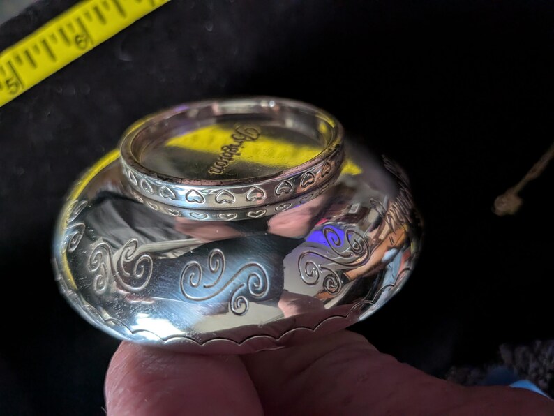 May include: A silver-toned metal bowl with a polished finish and engraved swirl patterns along the sides. A matching silver-toned ring with a heart-patterned band and the word Brighton engraved on the inner rim rests on top of the bowl.