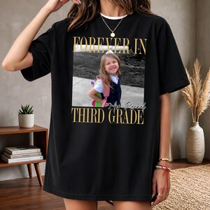Może przedstawiać: Czarny t-shirt z krótkim rękawem z centralną prostokątną grafiką przedstawiającą zdjęcie małego dziecka z plecakiem. Napis FOREVER IN THIRD GRADE oraz Mrs. Rachel jest nadrukowany złotymi literami nad i pod fotografią.