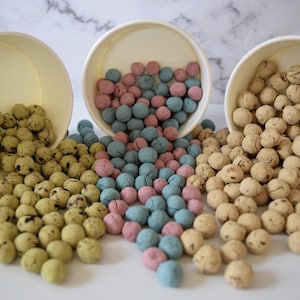 May include: Three white paper bowls tilted forward, each filled with small, round, textured spheres. The left bowl contains light green spheres, the center bowl holds a mix of pink and blue spheres, and the right bowl contains tan spheres.