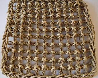 May include: A square-shaped mat constructed from woven natural tan fibers. The mat features a grid-like pattern created by interlacing thick, twisted strands of dried grass, forming a series of open, uniform square gaps throughout the entire surface.