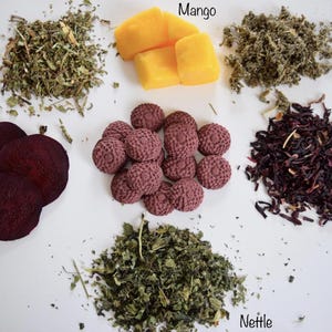 May include: A collection of ingredients including dried dandelion leaf, yellow mango cubes, dried raspberry leaf, dark purple beet slices, small round textured purple discs, dark red hibiscus petals, and dried green nettle leaves.