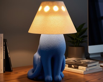 May include: A table lamp shaped like a sitting cat with a textured blue body and a conical off-white lampshade. The shade features a row of circular cutouts that emit light, creating a warm glow against the blue base of the lamp.