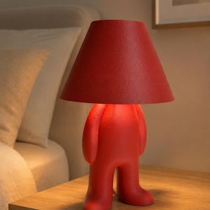 May include: A bright red table lamp shaped like a stylized human figure with a conical lampshade. The lamp features a smooth, matte finish on the base and a textured surface on the shade, emitting a warm glow from beneath the shade.