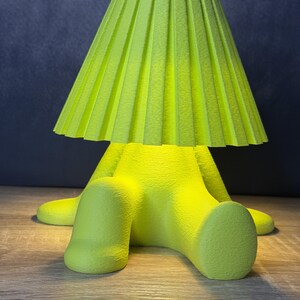 May include: A bright neon yellow table lamp featuring a seated figure base with a textured surface and a wide, pleated, conical lampshade. The lamp emits a soft, warm glow from beneath the shade, illuminating the figure's legs and the surface.