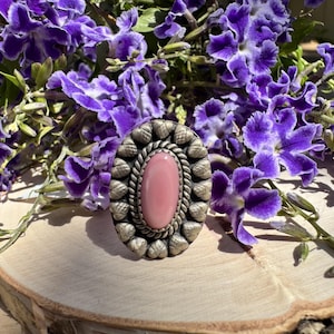 May include: An oval ring featuring a smooth, light pink cabochon stone at the center, surrounded by a twisted metal rope border and an outer edge of small, textured silver-toned metal acorn shapes arranged in a repeating pattern.