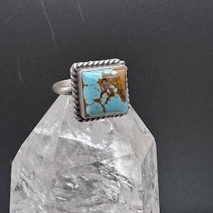 May include: A square ring featuring a light blue turquoise stone with brown matrix patterns, set in a silver frame with a twisted rope border. The ring band is smooth and silver, showcasing a polished finish on the metal setting.