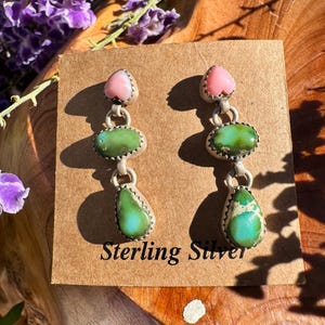 May include: Dangle earrings featuring a pink heart-shaped stone at the top, followed by an oval green stone and a teardrop-shaped green stone, all set in sterling silver. The earrings are displayed on a brown card with the text Sterling Silver.