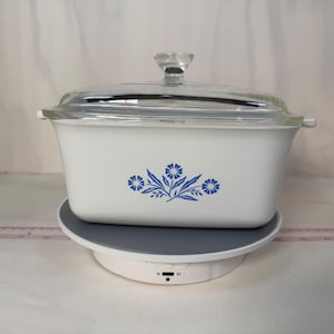 May include: A rectangular white casserole dish with a clear glass lid and a blue floral pattern on the front. The dish features small side handles and is positioned on a circular rotating display stand with a visible measuring tape nearby.