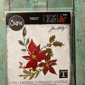 May include: A package of six die-cut metal dies for crafting. The dies feature a poinsettia flower with leaves and berries. The package is labeled "Sizzix Thinlits" and "Jim Holtz".