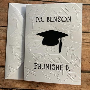 May include: White card with a black graduation cap and the text "DR. BENSON PH.D. FINISHED" on a background of embossed graduation caps.