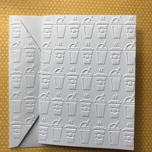May include: A white greeting card and matching envelope featuring an embossed, repeating pattern of coffee cups with lids and straws. The design consists of multiple rows of identical beverage containers, creating a textured, monochromatic surface.