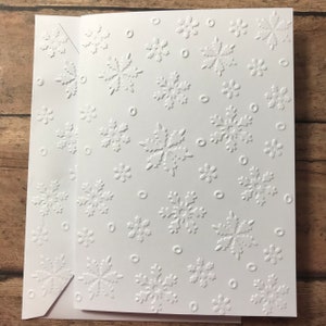 May include: White greeting card and envelope set with embossed snowflake pattern. The card features various sizes of snowflakes and small circles. The envelope has a matching snowflake design on the flap.