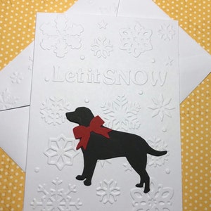 May include: White greeting card with embossed snowflakes and the phrase "Let it SNOW." A black dog with a red bow is featured on the card. Includes a white envelope. The background is yellow with white polka dots.